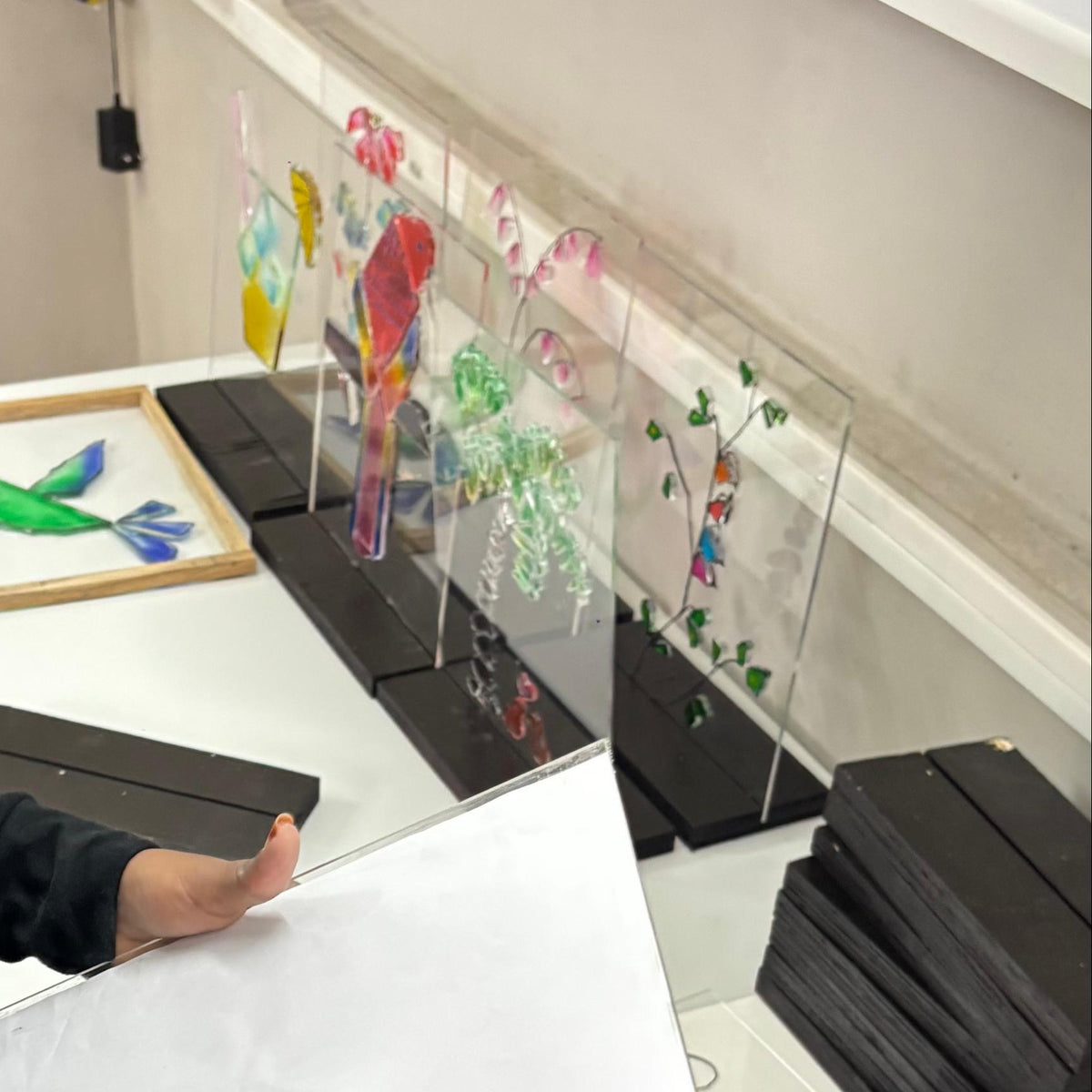 A person in a black long-sleeve shirt stands at a white desk, holding a sheet of white paper and placing a small colorful flower-shaped object on it. The desk also has a gray tape dispenser, black stacked items, and colorful hanging craft displays against a light wall.