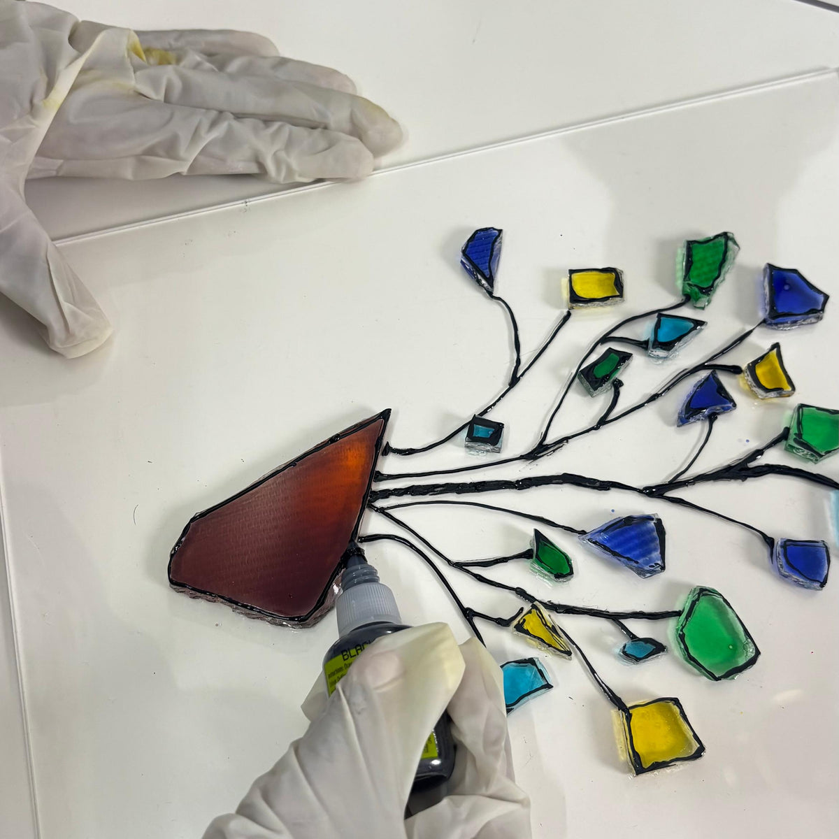 A person wearing white gloves is applying a piece of stained glass to a white panel. The panel features a black wire branch design with multiple colorful glass pieces in blue, green, yellow, and a reddish-brown piece being attached.