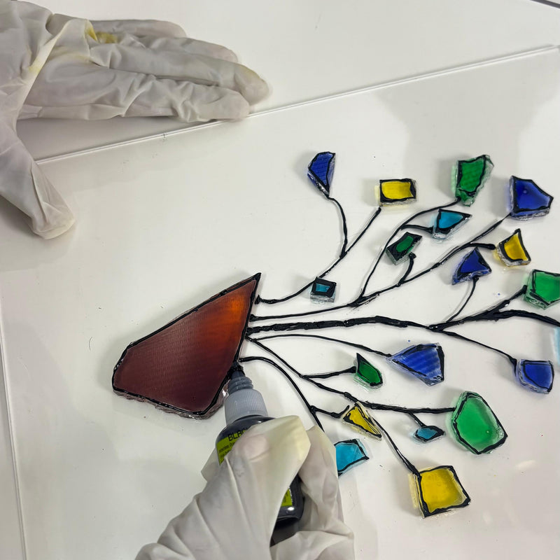 A person wearing white gloves is applying a piece of stained glass to a white panel. The panel features a black wire branch design with multiple colorful glass pieces in blue, green, yellow, and a reddish-brown piece being attached.